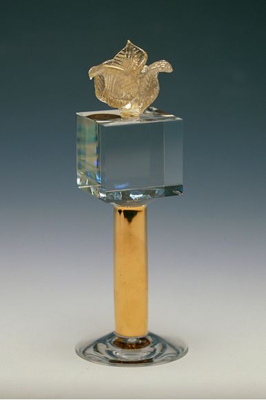 Titel: Porträtt av min Dotter: Du är min Rosenknopp Material: Kuben är av slipat och polerat optiskt glas, Rosen är gjord på venetianskt vis, runt det varma glaset har jag lagt på 24K bladguld, Det gyllene benet är målat med en guldhaltig vätska som benämns Glansguld för att guldet glänser efter bränning i en ugn. (Finns även som Polerguld, men denna måste poleras med en agat-sten efter bränningen.) Rosen är limmad på kuben som är limmad på ben och fot. Design och utförande: Anna Carlgren Fotograf: Anna Carlgren Höjd: 18 cm